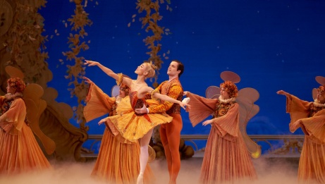 One dancer holds another wearing a tutu as she leans forward with arms extended on pointe. There are other dancers in the background wearing angel wings. 