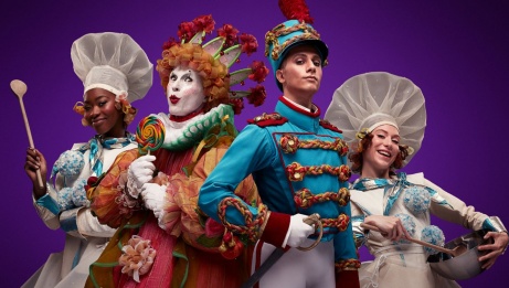 Four people post in front of a red backdrop. The outside two are dressed as chefs, holding wooden spoons. The person on the center-right is wearing a crown and holding a lollipop, and the person on the center-left is dressed as the Nutcracker, holding a sword.
