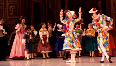 A scene with many things happening at once. A group of children stand off to the side in party clothes with a few adults, watching a pair of performers in geometric costumes, white hats, and rosy cheeks. 