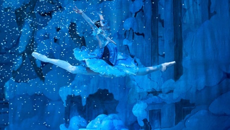 A dancer wearing a tutu leaps through the air in the splits with her arms above her head. There is a blue light and snow is falling. 
