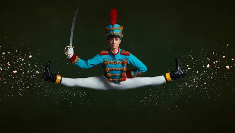 A dancer dressed as the Nutcracker holding a sword is in midair, in the splits, with glitter on the image, in front of a black background. 