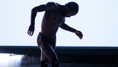 The silhouette of a dancer, in movement, arms in opposite direction of their body, in front of a bright projector screen. 