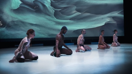 Five dancers, all bare-chested, kneel on stage in front of a projected photograph in undergarments. 