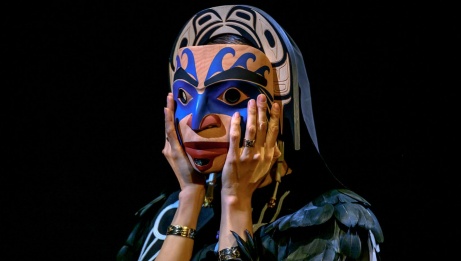 An Indigenous dancer in a raven winged cloak holds a mask to their face. 
