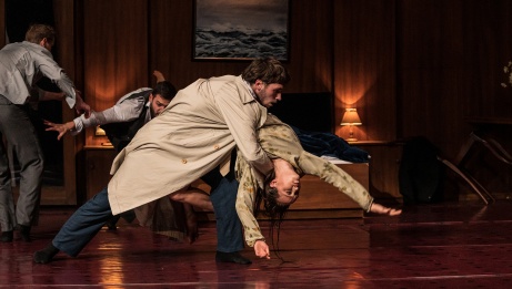 A dancer is caught in a dip by another dancer in a beige trench coat. Two other dancers are in motion behind them in a room lit by table lamps. 
