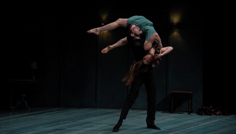 One dancer in a green dress is flung over the shoulder of another on a bare stage. 