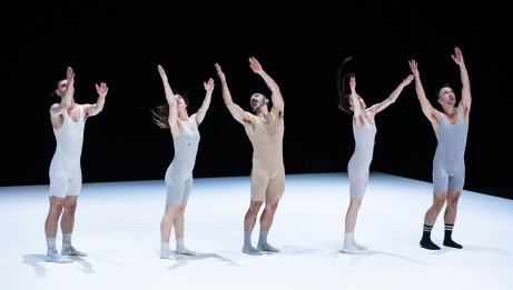 5 dancers in light toned unitards stand in a line with arms reaching upwards. 