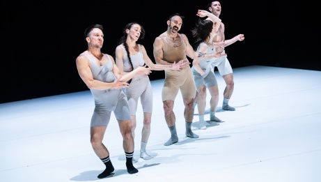 Five dancers in light toned unitards stand in a diagonal line with bent knees and hips to one side.