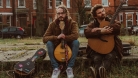 Deux hommes, un tenant une mandoline et l'autre une guitare, posent assis sur le trottoir devant un bâtiment en briques.  © Claire Loughran