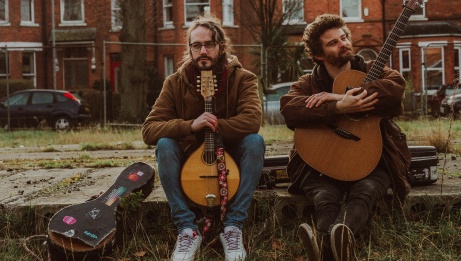 Deux hommes, un tenant une mandoline et l'autre une guitare, posent assis sur le trottoir devant un bâtiment en briques. 
