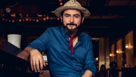 Le musicien est assis de manière décontractée dans un bar, portant un chapeau, une chemise bleue et un petit foulard rouge autour du cou. © Quique Escamilla