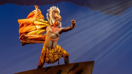 Simba stands proudly at the edge of Pride Rock. A lion mask made of wood sits atop his head as his clothes flow like a lion’s mane in the wind. © Disney Photo by Deen van Meer