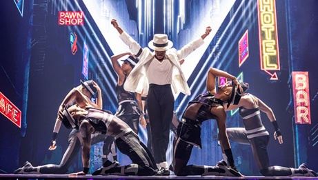 MJ se tient debout au centre de la scène, les bras tendus vers le ciel, dans son célèbre costume blanc. Il interprète en compagnie d’autres artistes la chanson Smooth Criminal. © Roman Banks dans le rôle de MJ et la distribution de la première tournée nationale.