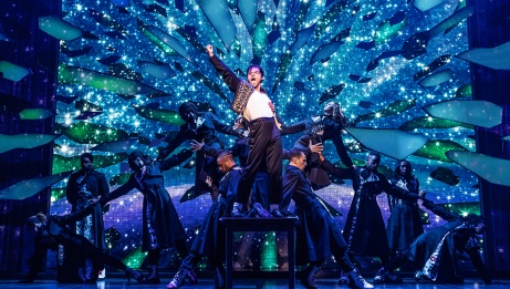 MJ stands on a table with one fist in the air. A group of dancers form a human link behind him. Broken shards of glass float behind them.