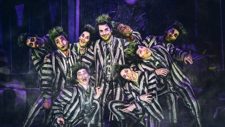Beetlejuice stands in the centre of a group, dressed in his signature black and white suit and green spiky hair. Company members surround him, dressed in identical suits, making various silly faces.
