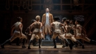 Hamilton stands center on a platform looking upward with a single spotlight overhead. A group of dancers surround and lean towards him. © Joan Marcus, 2021