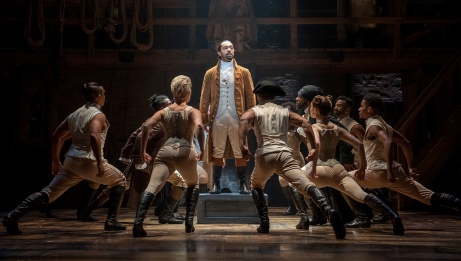 Hamilton stands center on a platform looking upward with a single spotlight overhead. A group of dancers surround and lean towards him.