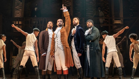Hamilton stands center and points to the sky as his friends stand beside him. There are company members on either side of them with their fists upwards.