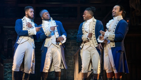 Hamilton and friends, representing different backgrounds, are dressed in blue colonial clothing. The two on the left point to the laughing men on the right.