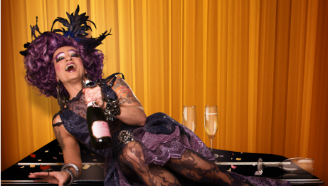 Mado Lamotte, allongée sur un piano, riant et tenant une bouteille de champagne