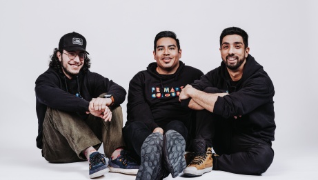 The 3 band members sitting on the floor in front of a white background.