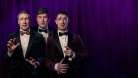 The 3 comedians wearing suits and bowties, in front of a dark blue curtain, making funny faces. 