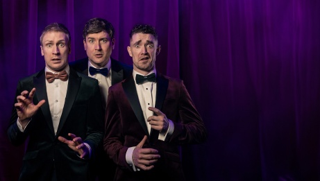 The 3 comedians wearing suits and bowties, in front of a dark blue curtain, making funny faces.