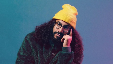 Apollo the Child is wearing a yellow beanie, dark-rimmed glasses, and a green fur jacket. He is posing with his head resting on one hand and looking directly at the camera against a gradient blue and purple background.