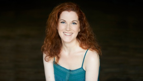 Genna smiling and looking at the camera. She's wearing a turquoise tank top and she has red curly hair.