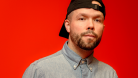 D-Track standing in front of a red background and wearing a backwards cap. © Scott Doubt