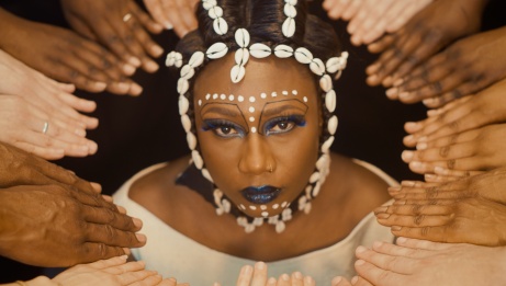 Djely Tapa surrounded by numerous hands is looking up at the camera while wearing a crown of shells.