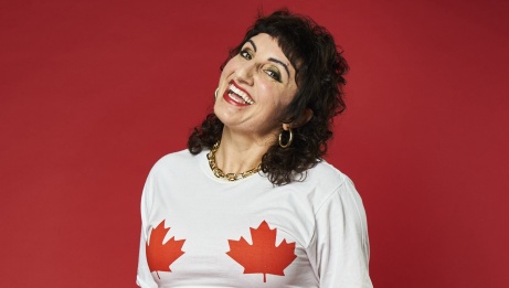 Steph sourit, elle porte un t-shirt blanc avec des feuilles d'érable et elle est devant un mur rouge.