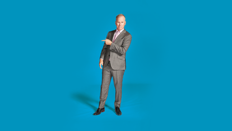 Gerry Dee wearing a suit and pointing at his right. He's in front of a blue background.