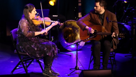 Performers on stage playing the fiddle and guitar.