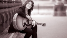 Photo sépia de Rosanne Cash. Elle est assise dans la rue avec sa guitare. © Pamela Springsteen