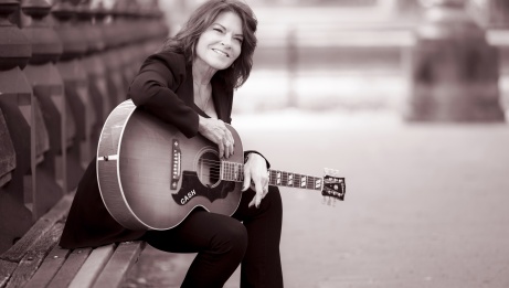 Sepia photo of Rosanne Cash. She is sitting in the street with her guitar. © Pamela Springsteen