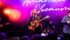 Amanda Rheaume is singing and playing the guitar on stage. There is pink smoke behind her.  