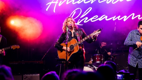 Amanda Rheaume is singing and playing the guitar on stage. There is pink smoke behind her.  