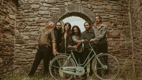 The members of El Balcón standing in a stone archway and holding a bike. 