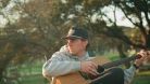 Dylan Gossett sitting outside on a beautiful day and there are trees behind him. He's wearing a grey hoodie and a cap, and he's playing acoustic guitar.  