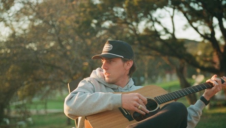 Dylan Gossett sitting outside on a beautiful day and there are trees behind him. He's wearing a grey hoodie and a cap, and he's playing acoustic guitar. 