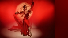 A ballet dancer in a flowing red dress stands en pointe against a red background. She holds a sheer tulle fabric with one arm extended above her head, and the other to the side while a yellow flare of light adds a dramatic glow to the scene. © Danny Shumov