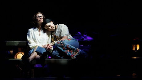 A White mother seated and her Métis daughter cuddled next to her. The mother wears a white shirt and jeans, and the daughter wears a white shirt, beaded denim pants, and her hair in braids. 
