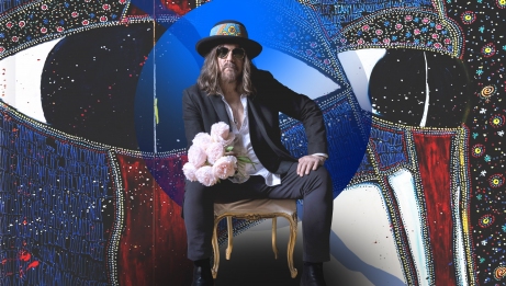 Tom Wilson sits on a chair holding a bouquet of flowers, with one of his paintings in the background.
