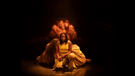 Three women on stage: one crouching at centre facing the audience and looking to the left, the other two leaning against her back on either side. The reflection of the women behind appears in a mirror at the rear.