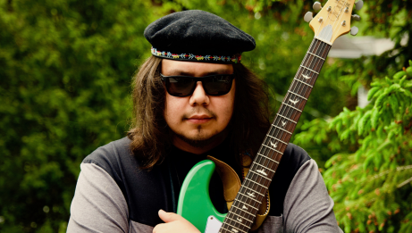 A person wearing a beret and sunglasses stands outdoors holding a bright green electric guitar.