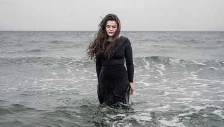 Une personne v&ecirc;tue d&rsquo;un haut sombre &agrave; manches longues se tient debout dans l&rsquo;oc&eacute;an, face &agrave; l&rsquo;eau. Elle a de l&rsquo;eau jusqu&rsquo;&agrave; la taille. Ses longs cheveux sont souffl&eacute;s sur le c&ocirc;t&eacute; par le vent, et les vagues ondulent autour d&rsquo;elle sous un ciel mena&ccedil;ant. 