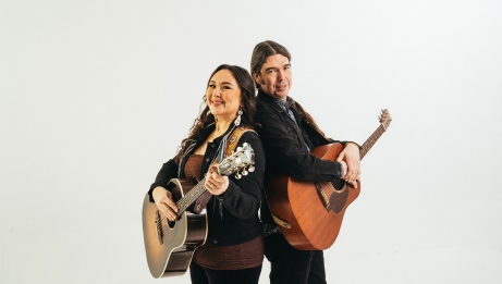 Two individuals stand back-to-back holding acoustic guitars. Both wear dark clothing, and the background is a clean white studio setting.