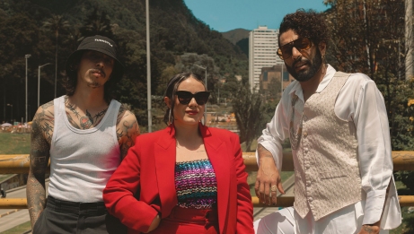 Trois artistes posent &agrave; l&rsquo;ext&eacute;rieur devant une ville et des collines verdoyantes, portant des tenues audacieuses et des lunettes de soleil. 