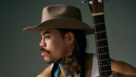Close-up profile of a person wearing a wide-brim hat, with braided hair and visible neck tattoos, holding an acoustic guitar against a plain background.